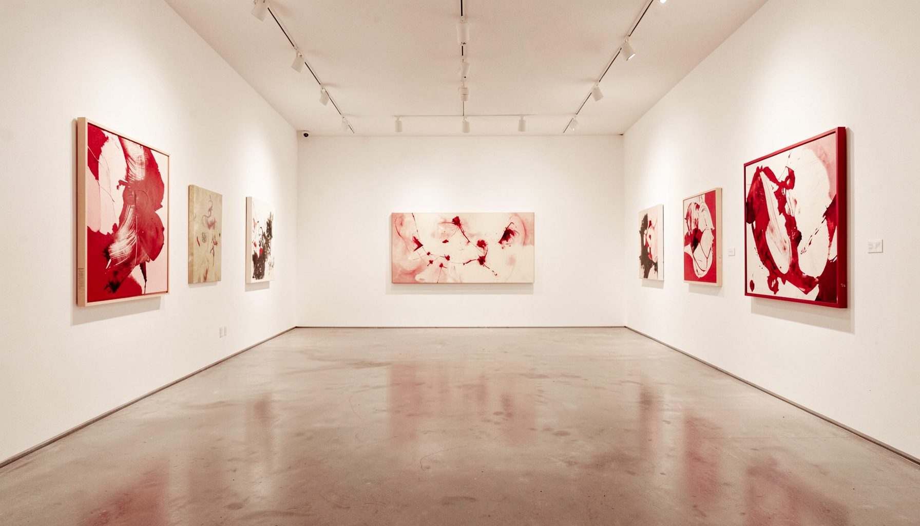 Un interno di galleria d'arte contemporanea con pareti bianche illuminate, pavimento in cemento chiaro e opere astratte appese; dominano toni caldi di bianco, crema e tocchi di rosso acceso.