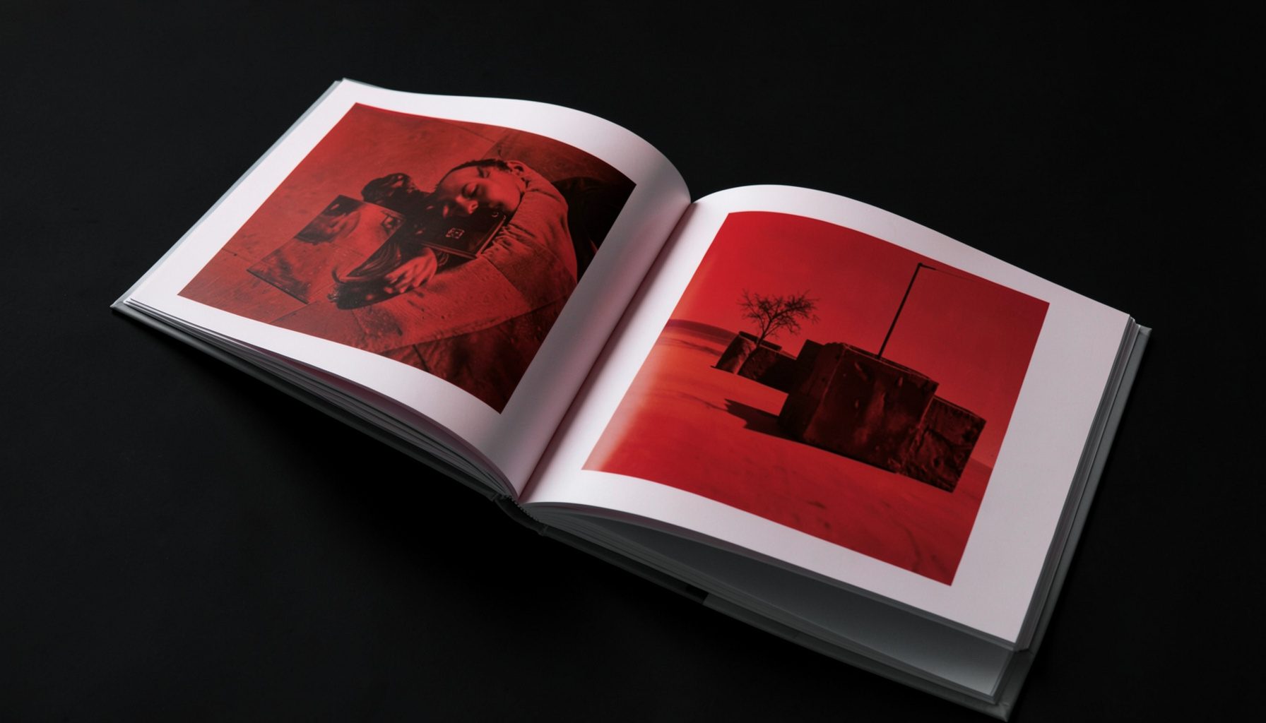 Primo piano di un catalogo d'arte rilegato aperto su una superficie scura, pagine lucide con riproduzioni di opere contemporanee dai toni caldi del rosso e dell'ocra, illuminazione soffusa laterale.