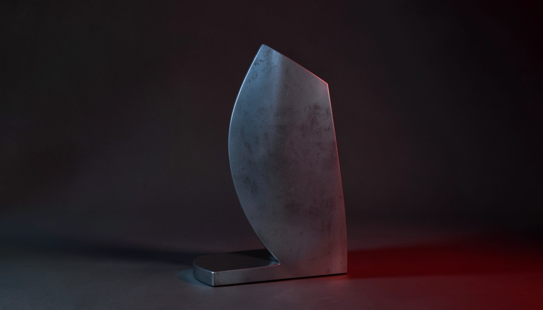 Scultura in metallo dalla silhouette essenziale, fotografata in controluce con toni grigio-argento e blu, resa in duotono.
